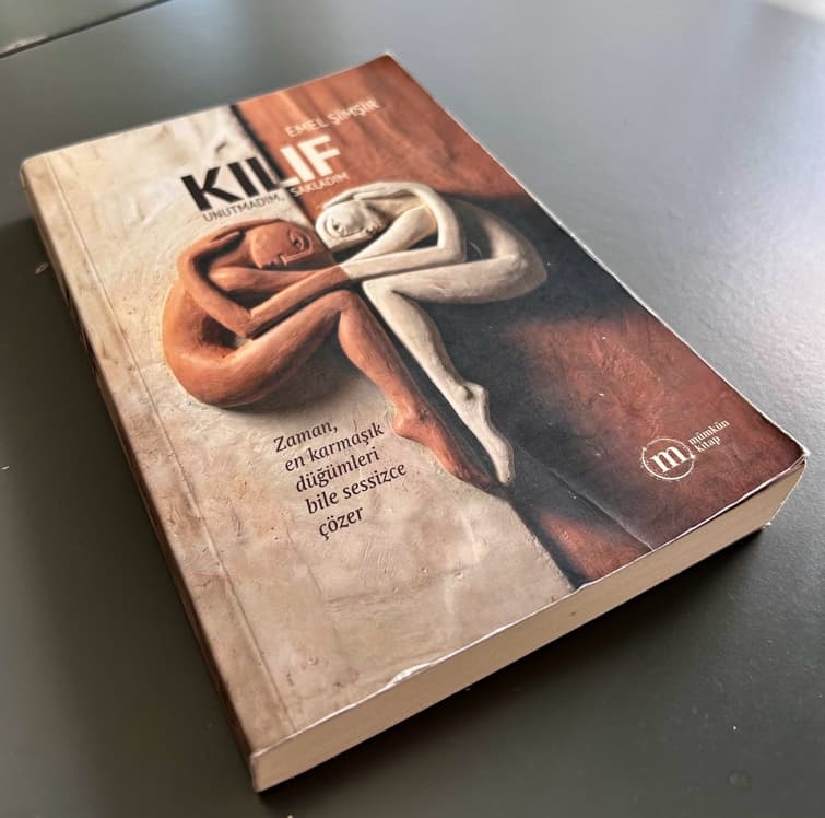 Masanın üzerinde duran bir Kılıf romanı; kapakta kil rengi ve beyaz, birbirine sarılmış iki figür.